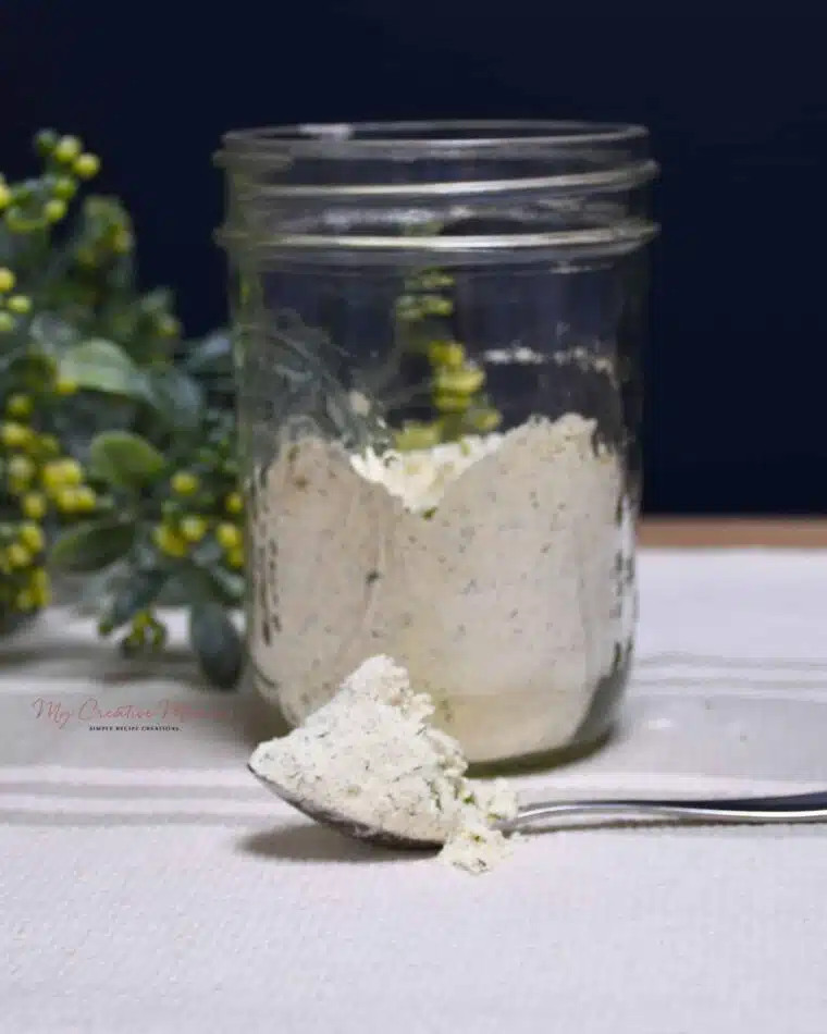 A spoon that has some of the homemade ranch seasoning recipe on it with a mason jar that is filled with more of it behind the spoon.