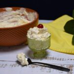 The front view of a dish that has pineapple dreamy salad in it with a cherry on top. Behind the dish is a large bowl that is filled with the salad.