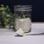 A mason jar that is half filled with the homemade ranch seasoning recipe with a spoon that is in front of the jar with more of the blend on it.