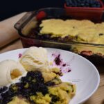 Close up of a bowl filled with the blueberry lemon cobbler with more of the 3 ingredient dessert in a baking dish behind the bowl.