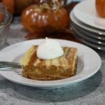 A close up of a white plate that has a pumpkin cheesecake bar on it with a dollop of whipped topping on it.