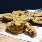 Close up of a stack of the Oreco chocolate chip cookies with more on a wire cooling rack behind the stack.