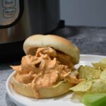 Creamy buffalos chicken on a bun with chips next to it and an Instant Pot in the background.