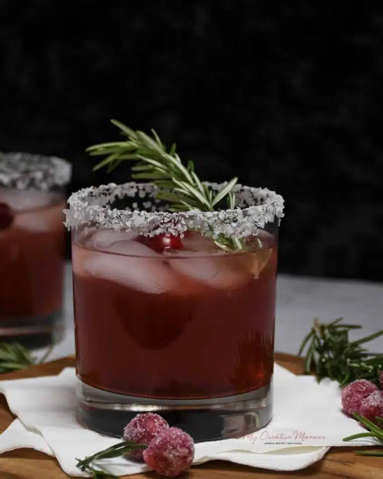Close up of a cocktail glass that has a salted rim and filled with a cranberry margarita. Garnished with a sprig of rosemary and fresh cranberries.