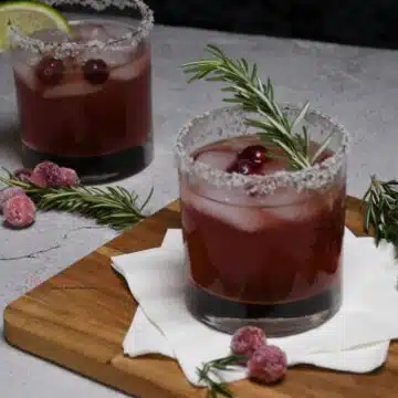 Two cocktail glasses that are filled with the cranberry margarita and garnished with a lime wedge, fresh cranberries, and a rosemary sprig.