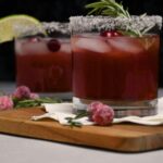 Close up of a cocktail glass that has a salted rim and garnished with fresh cranberries and a sprig of rosemary.