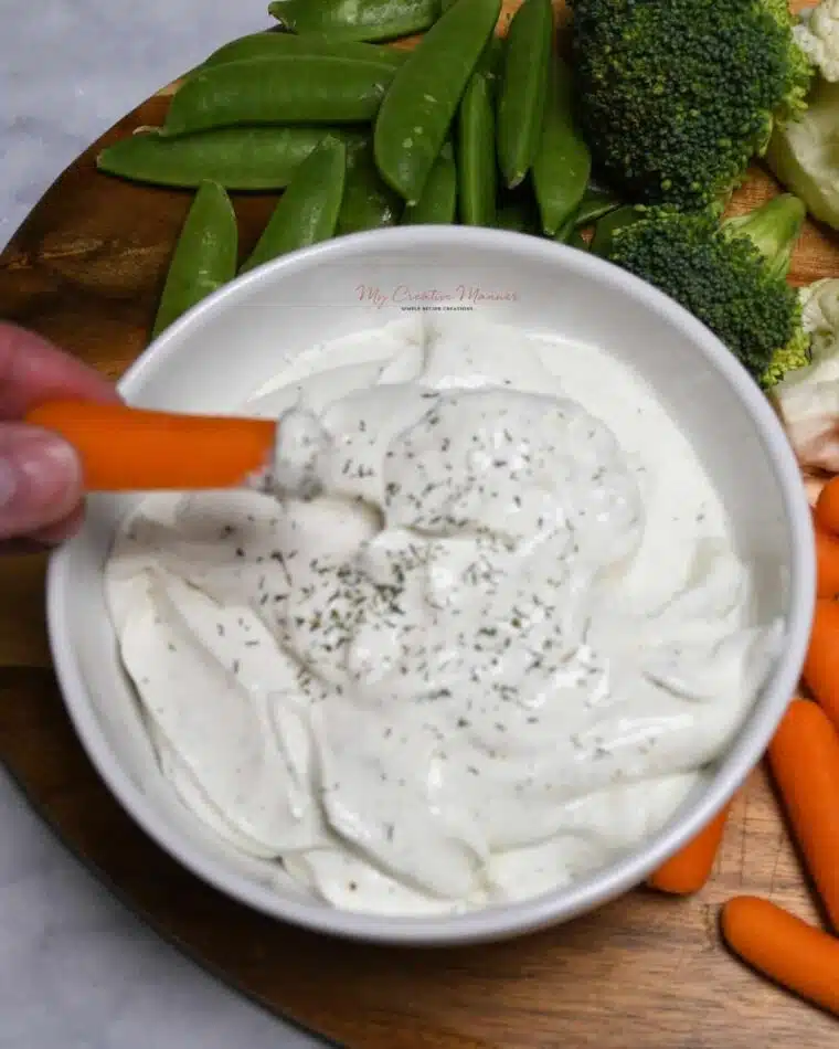 A hand that has a carrot that has been dipped into a bowl of ranch cottage cheese dip.
