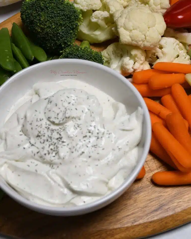 A bowl filled with creamy cottage cheese ranch dip that's on a platter with veggies around the bowl.