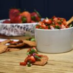 A pita chip that has the strwberry salsa on it with more in a bowl behind the chip.