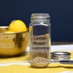 The front of a spice jar with a measuring spoon in front of it, both have the homemade pepper lemon seasoning blend in them.