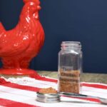 A measuring spoon and a spice jar filled with the homemade chicken taco seasoning recipe with a red chicken in the back ground.