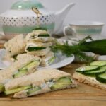 Half of a tea sandwich on a board with more of the cucumber cream sandwiches on a plate.