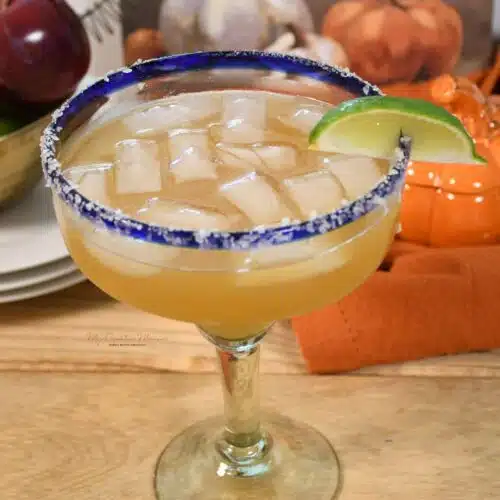 A glass filled with a apple cider margarita cocktail that is garnished with cinnamon salt rima nd a lime wedge.